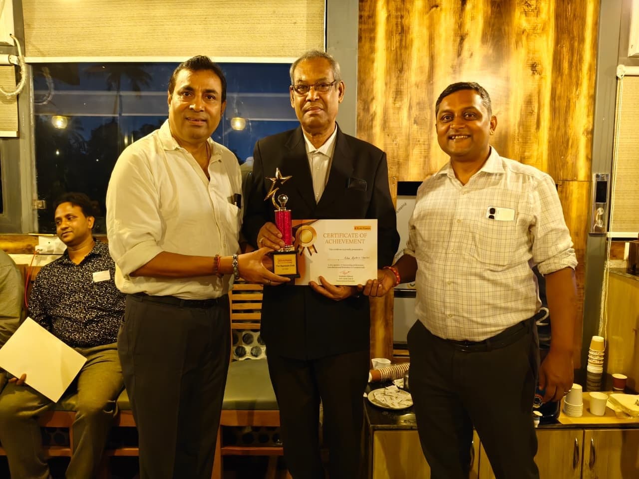 Three men standing together; the man in the centre holds a gold star trophy and a Certificate of Achievement